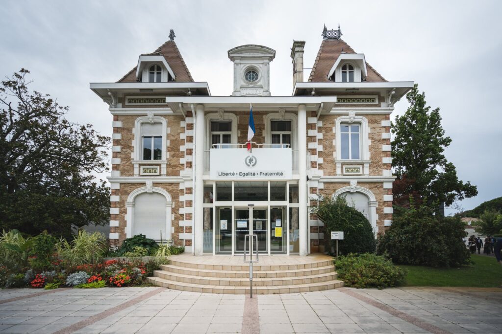 Façade en briques et pierre d'une mairie à tourelles, sous un ciel nuageux, avec des massifs de fleurs devant l'escalier.