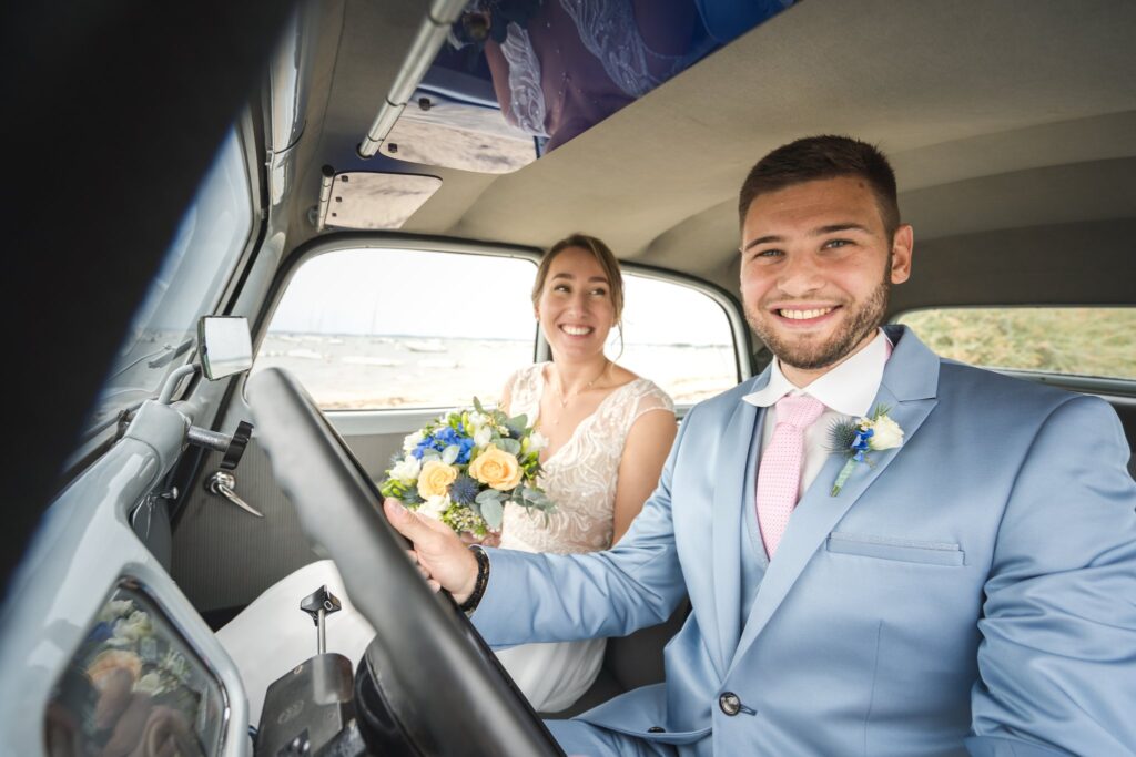 Un marié souriant au volant d'une voiture ancienne, avec la mariée et son bouquet assise à côté de lui.