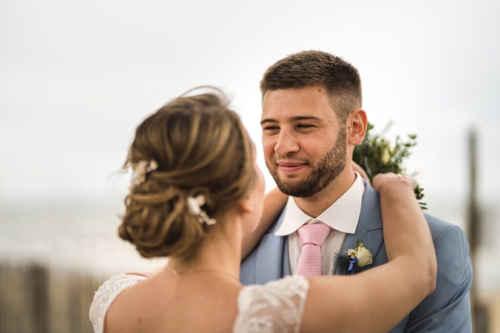 Une mariée de dos, le bras autour du cou du marié qui sourit, avec la mer floue en arrière-plan.