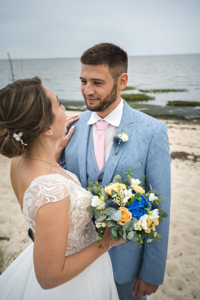 Sur une plage, la mariée regarde le marié en tenant un bouquet bleu et jaune, la mer derrière eux.