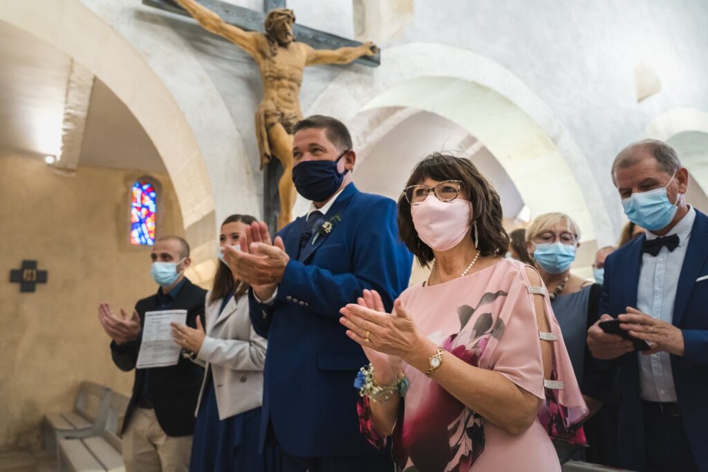 Des invités masqués applaudissent dans une église sous un grand crucifix de bois.