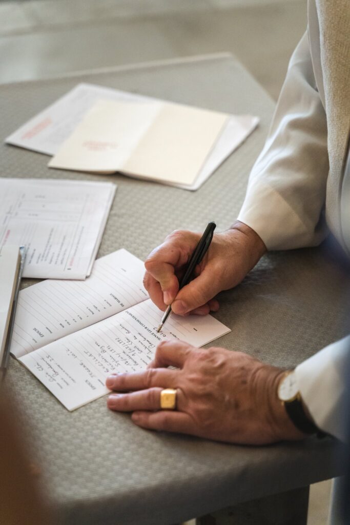 Une main tenant un stylo remplit un registre posé sur une table, avec des documents autour.