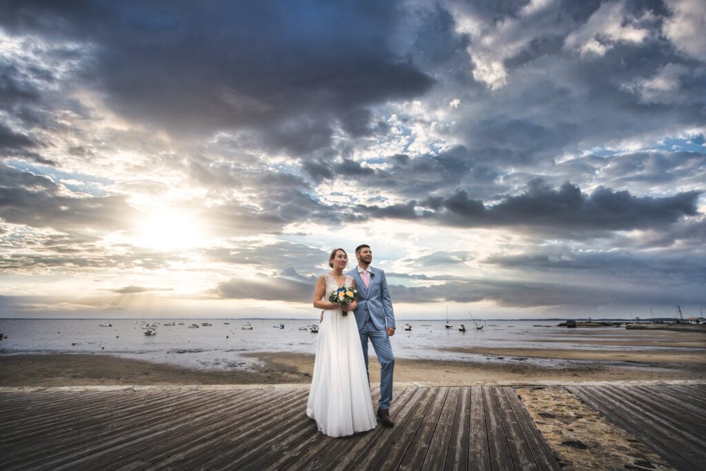 Les mariés au loin sur une plage à marée basse, sous un ciel de nuages sombres traversé par le soleil.