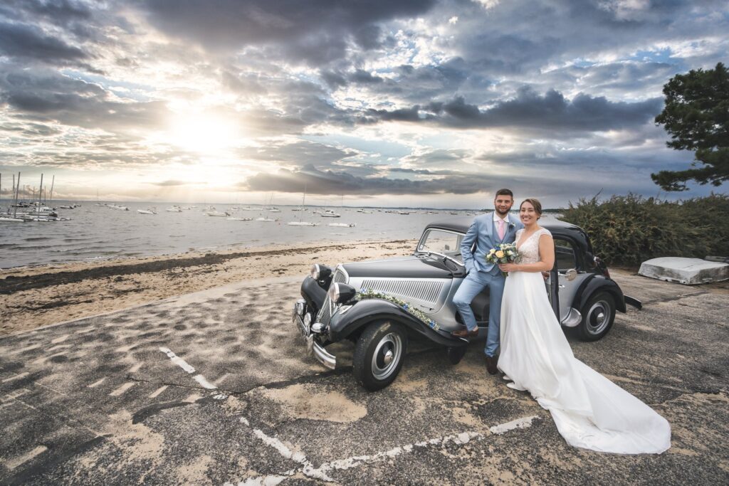 Les mariés posent sur une plage du bassin d'Arcachon, appuyés contre une Citroën Traction ancienne, sous un ciel de coucher de soleil.