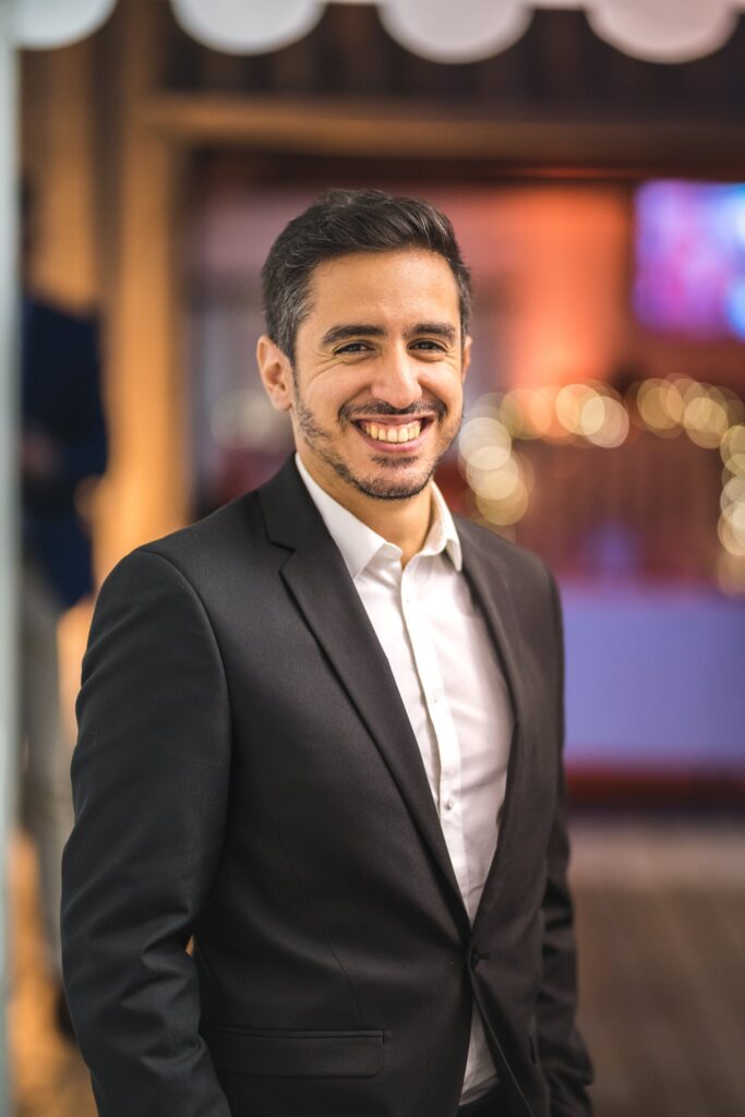 Portrait d'un homme souriant en costume sombre et chemise blanche, sur fond de lumières floues.