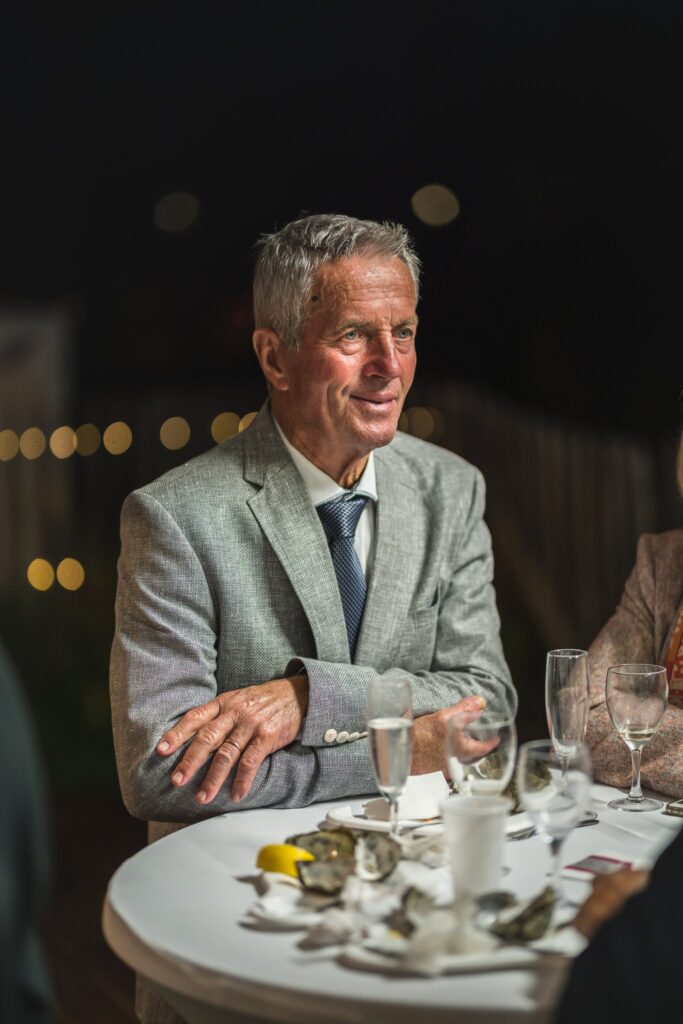 Un homme âgé en veste claire et cravate, assis à une table de nuit avec un plateau d'huîtres et des verres.