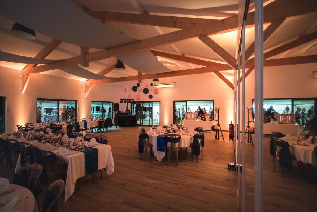 Vue large de la salle de réception éclairée en teintes chaudes, avec ses tables dressées et sa charpente en bois.
