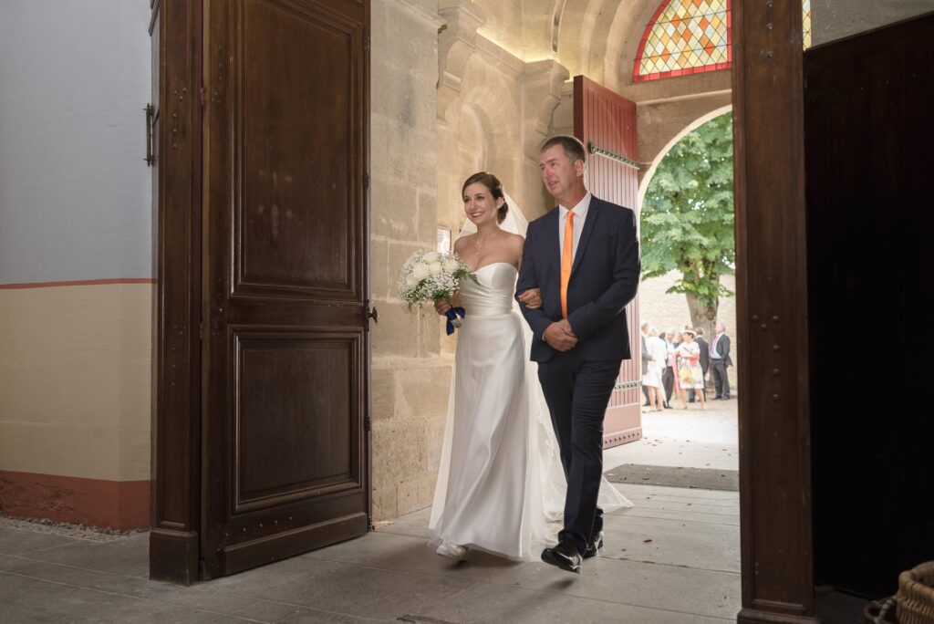 La mariée au bras d'un homme en costume franchit la porte en bois de l'église.