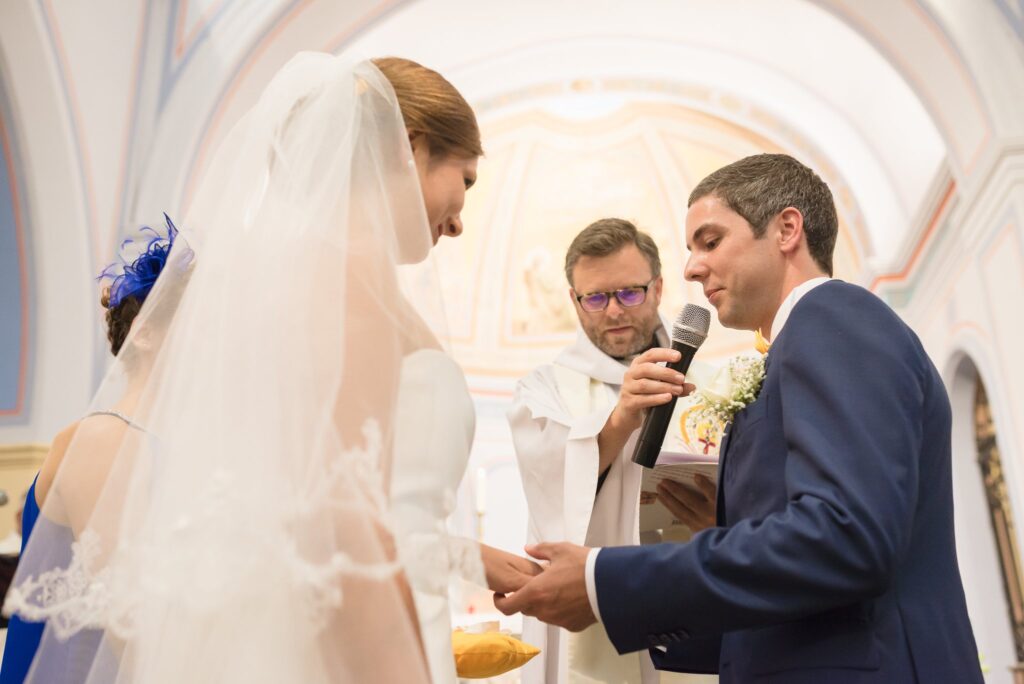 Le marié et la mariée voilée devant un prêtre qui tient un micro pendant la cérémonie.