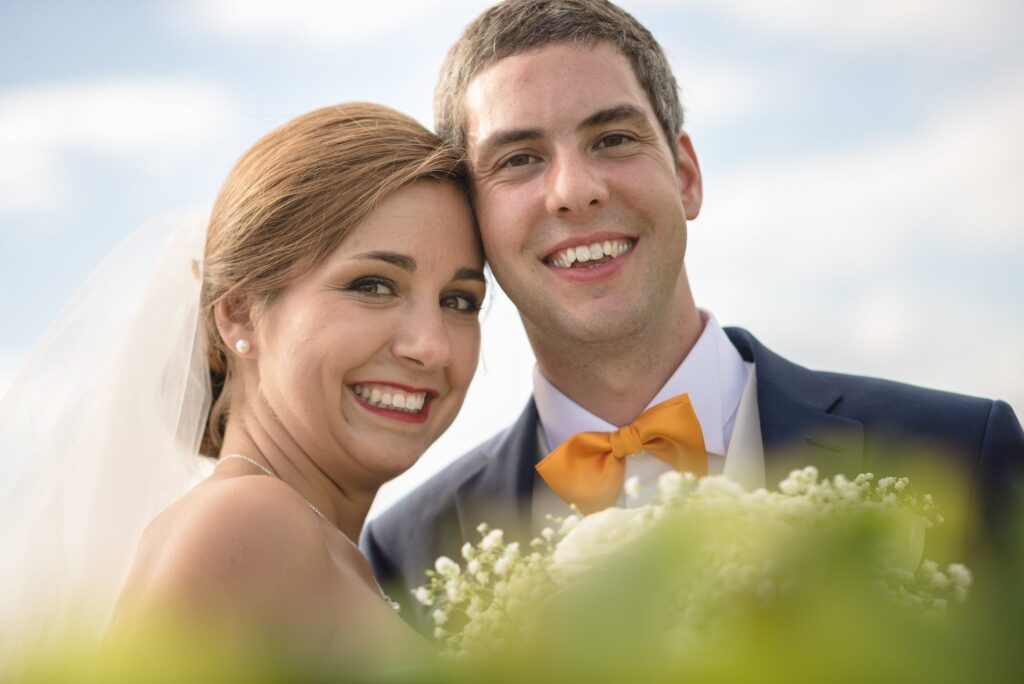 Portrait rapproché des mariés souriants, joue contre joue, avec un bouquet flou au premier plan.