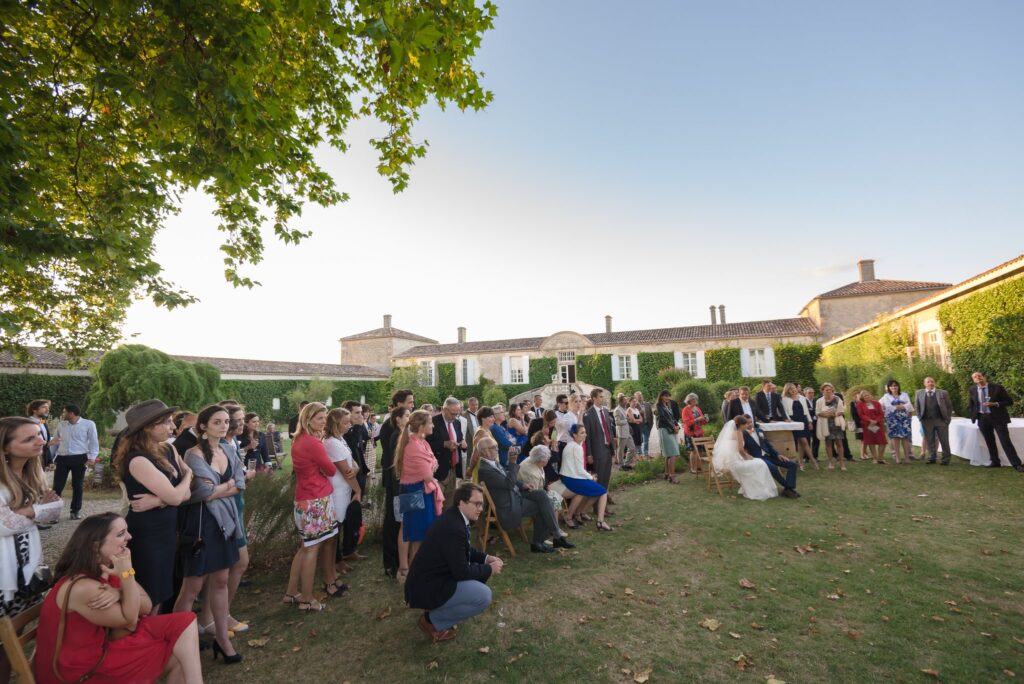 Les invités rassemblés sur la pelouse devant un long bâtiment, sous un grand arbre, en fin de journée.