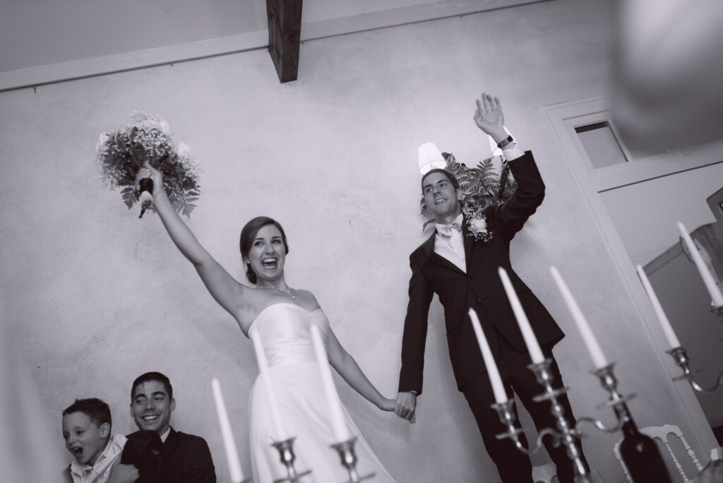 En noir et blanc, les mariés se tiennent par la main et lèvent le bras en entrant dans la salle.