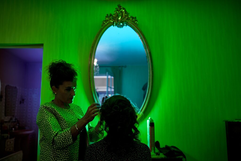 Une femme en chemisier vert à pois blancs coiffe une autre femme devant un grand miroir ovale, dans une pièce baignée d'une lumière verte.