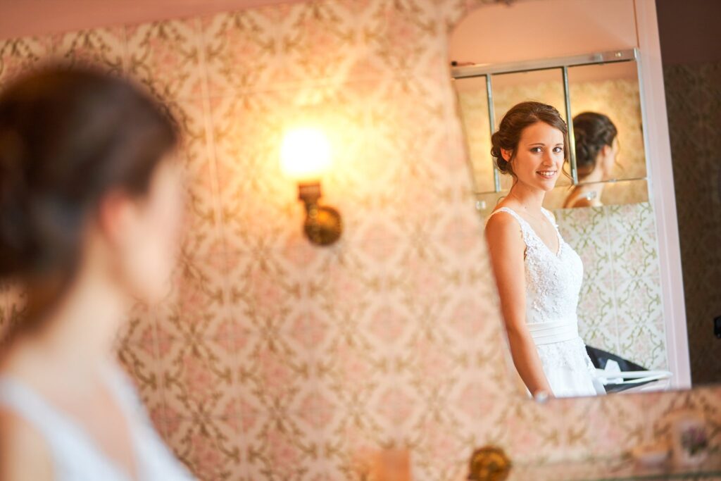 Reflet dans un miroir d'une mariée souriante en robe blanche, devant un mur à motif floral rose et blanc orné d'une applique dorée.