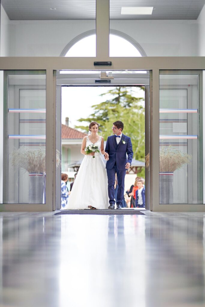 Un couple de mariés, la femme en robe blanche et l'homme en costume bleu marine, apparaît derrière les portes vitrées d'un bâtiment moderne.