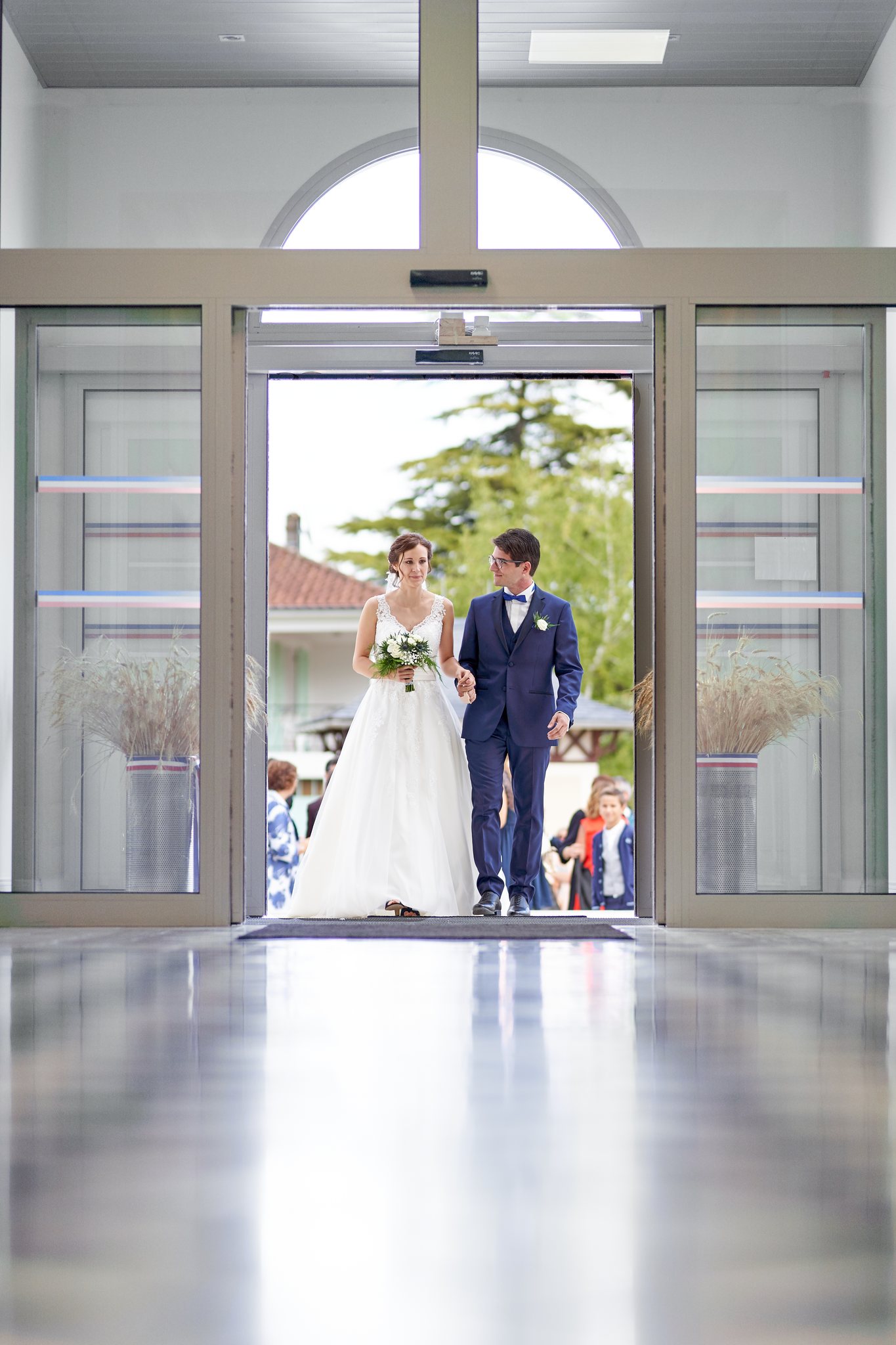 Un couple de mariés, la femme en robe blanche et l'homme en costume bleu marine, apparaît derrière les portes vitrées d'un bâtiment moderne.