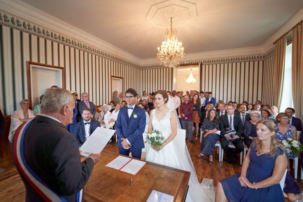 Cérémonie de mariage civile : le maire est assis devant les mariés, avec des invités assis et debout derrière eux, dans une salle aux murs rayés et au grand lustre.