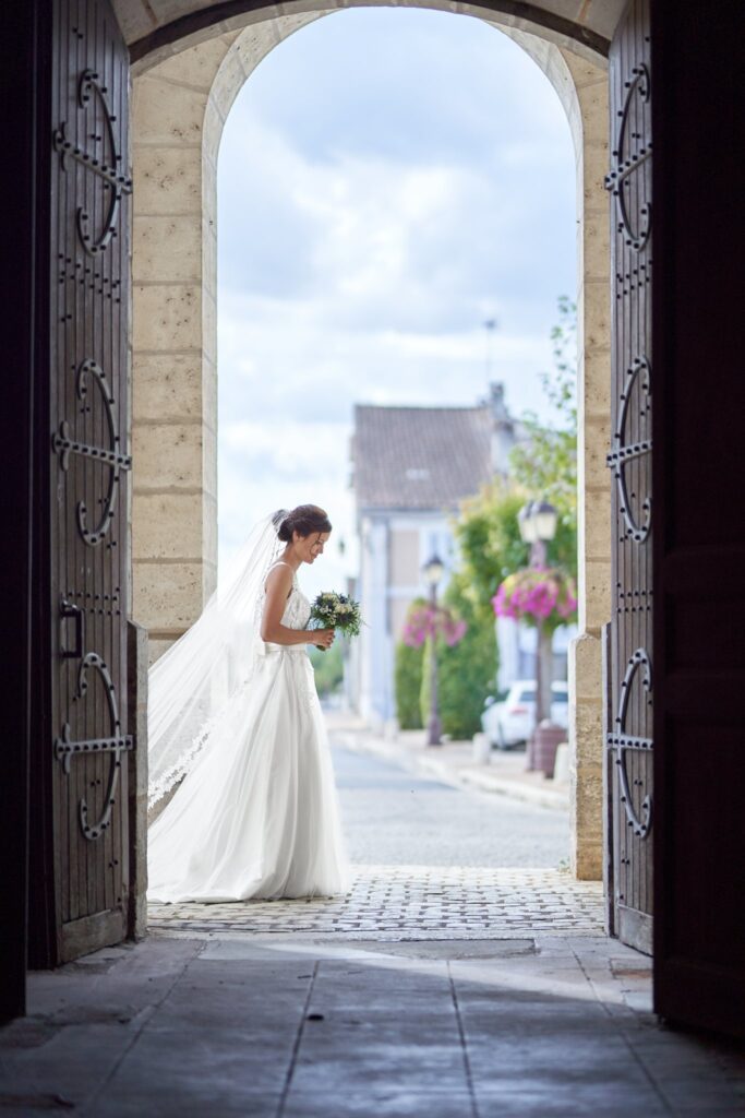 Une mariée en robe blanche et voile, un bouquet à la main, se tient dans l'embrasure d'une porte voûtée en pierre qui donne sur une rue de village sous un ciel nuageux.