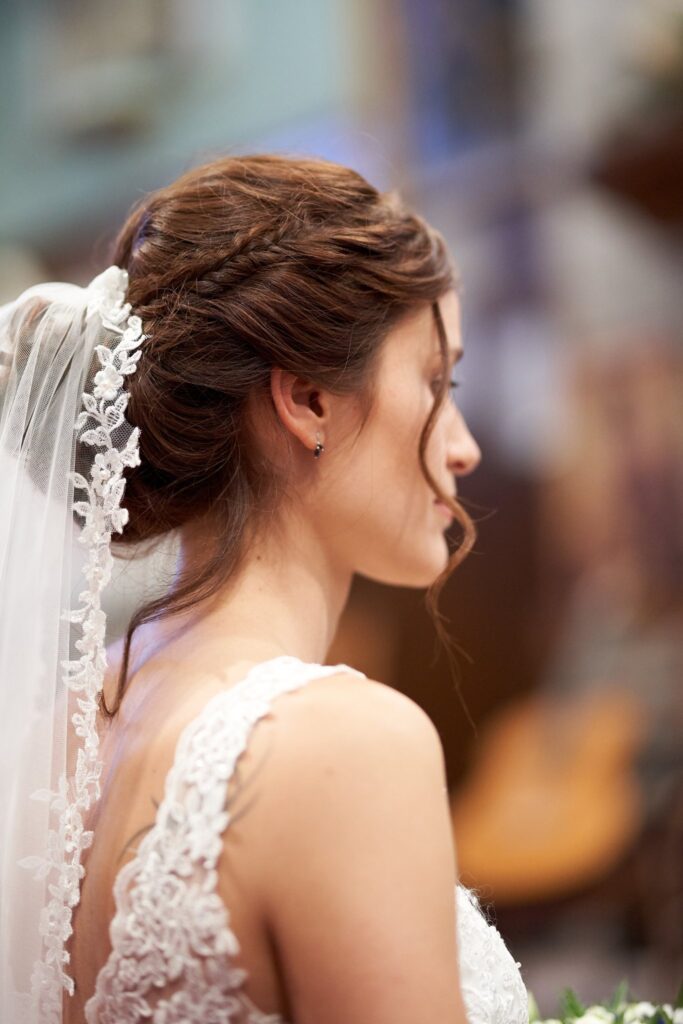 Portrait de profil d'une mariée aux cheveux bruns relevés, coiffée d'un voile en dentelle, en robe blanche à détails de dentelle.