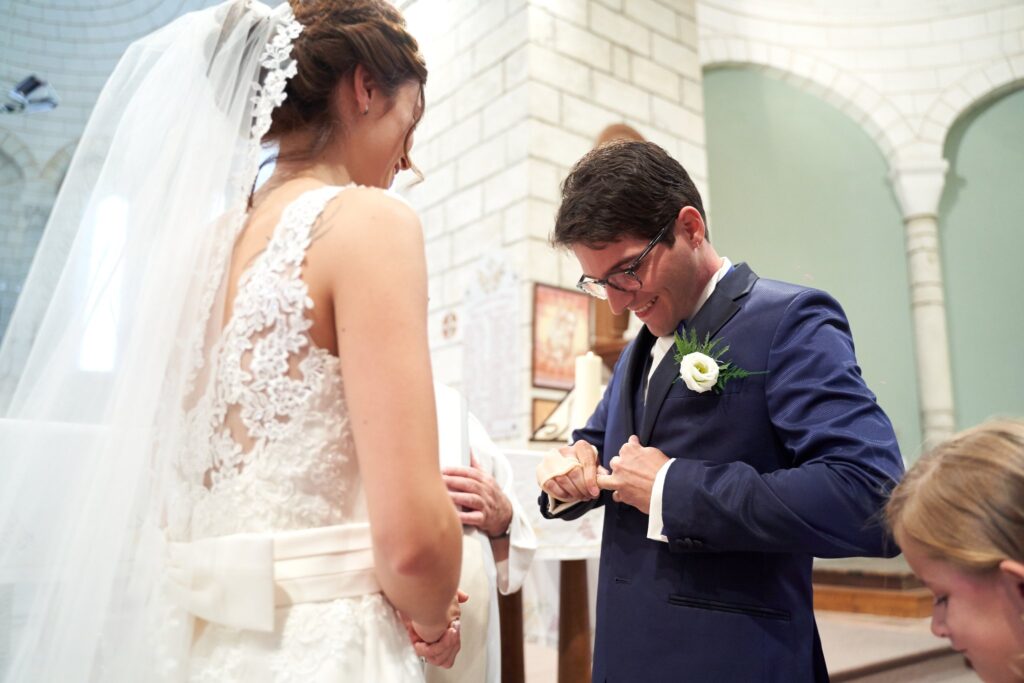 Un marié en costume bleu marine baisse les yeux vers ses mains, à côté d'une mariée en robe de dentelle et voile, dans une pièce aux murs clairs.
