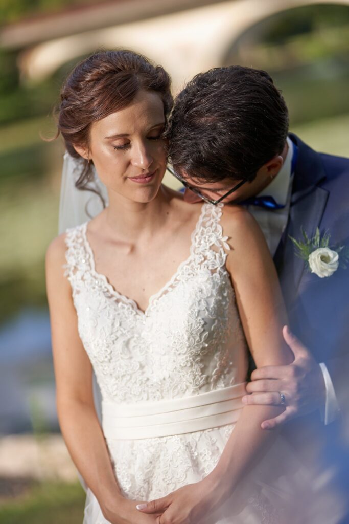 Le marié penche la tête contre le cou de la mariée, qui porte une robe blanche en dentelle, devant un fond de verdure flou.