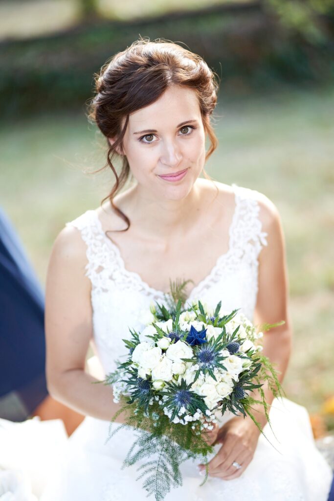 Portrait souriant d'une mariée en robe blanche en dentelle, tenant un bouquet rond de fleurs blanches et bleues, sur un fond flou.
