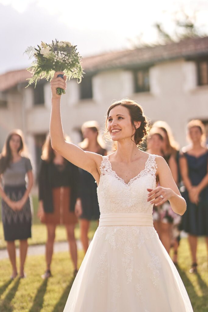 Une mariée souriante en robe blanche lève son bouquet de fleurs blanches pour le lancer à un groupe de femmes, devant des bâtiments blancs.