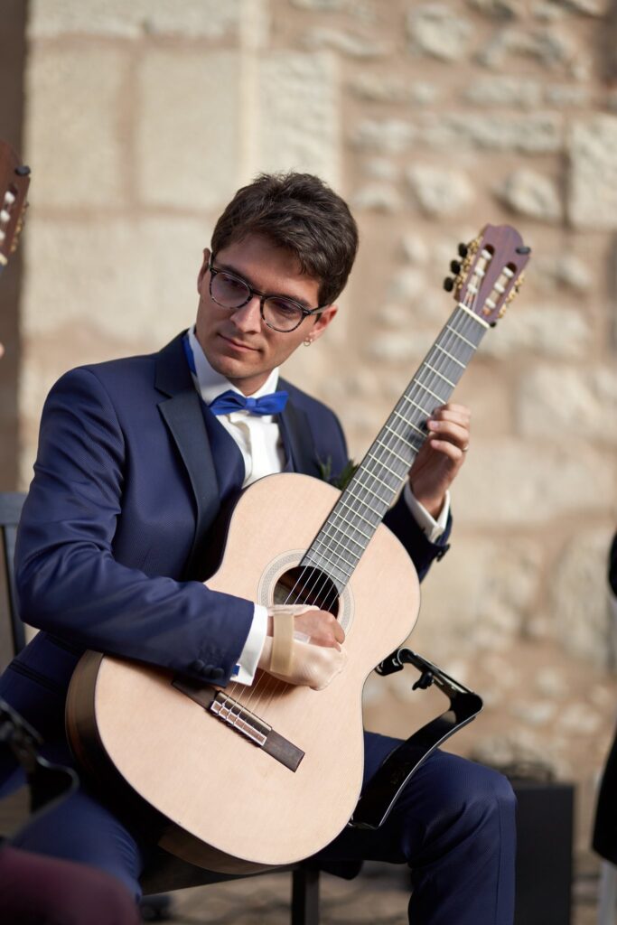Un jeune homme en costume bleu marine, lunettes et nœud papillon bleu, joue de la guitare classique devant un mur de pierre.