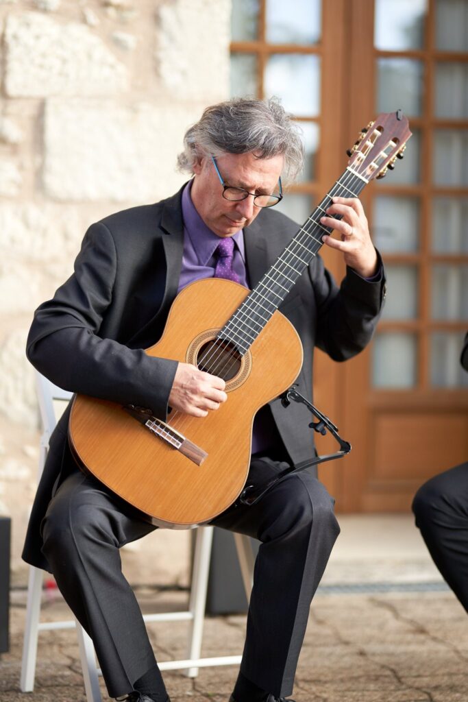 Un homme aux cheveux gris, en costume sombre et chemise violette, joue de la guitare classique, assis sur une chaise à l'extérieur.