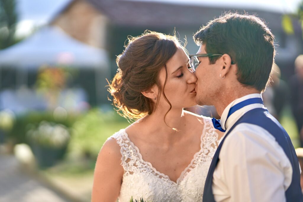 Gros plan d'un couple de mariés qui s'embrasse, la mariée en robe de dentelle blanche et le marié en costume bleu marine, devant un jardin flou.