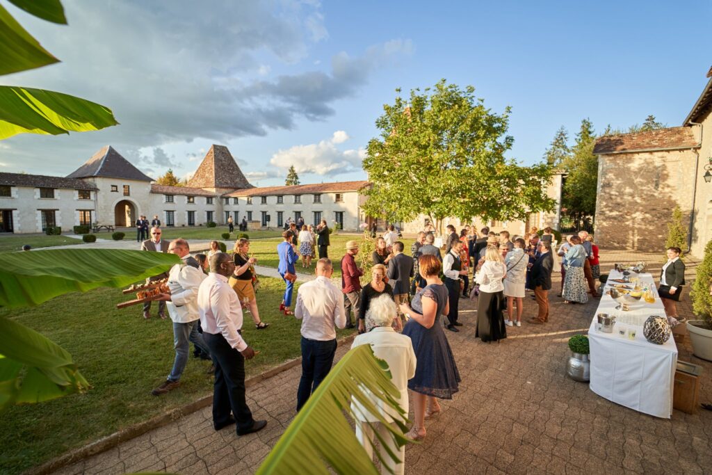 Des invités rassemblés sur une pelouse et une allée devant des bâtiments blancs, avec des arbres et une table de nourriture et de boissons.