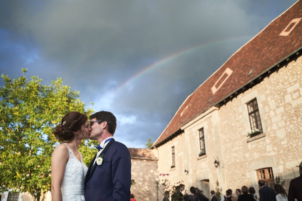 Un couple de mariés s'embrasse devant un bâtiment en pierre, sous un arc-en-ciel visible dans un ciel nuageux.