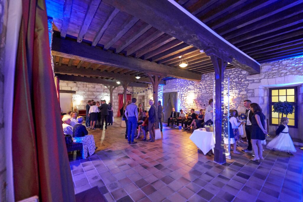 Une grande salle aux poutres apparentes et aux murs de pierre éclairés de bleu et de violet, où des invités sont réunis pour la soirée.