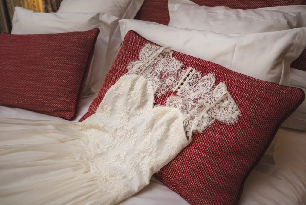 Une robe de mariée en dentelle posée sur un lit, entre des coussins rouges.