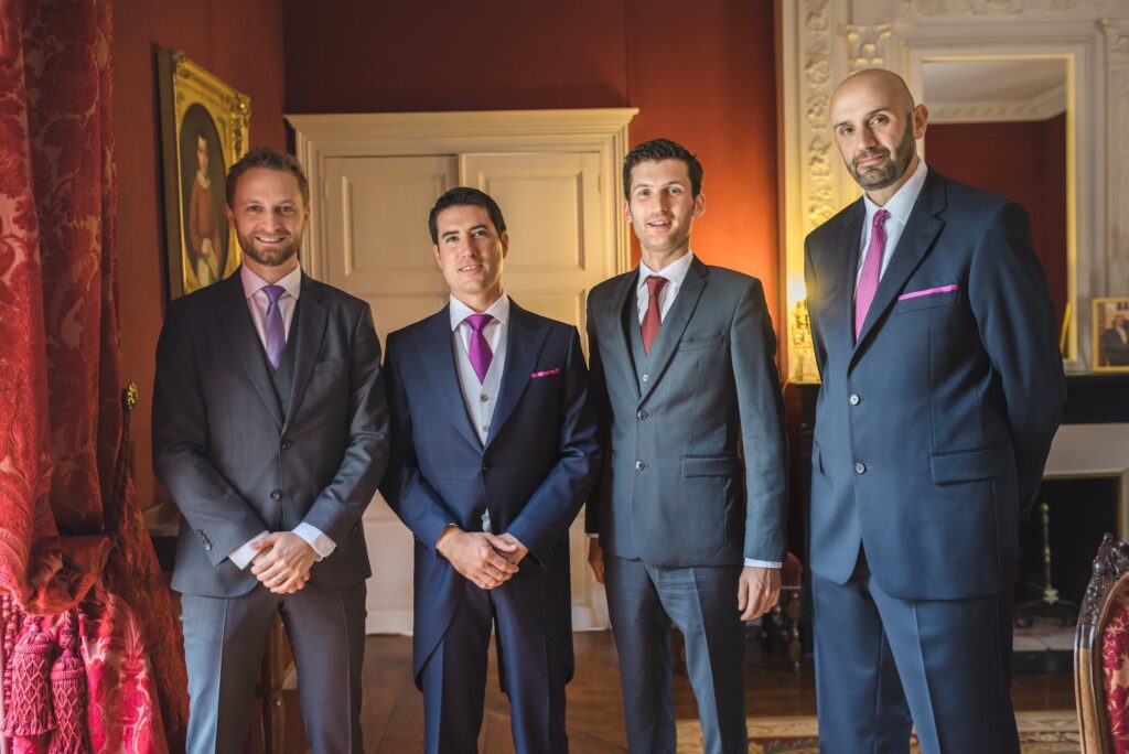 Quatre hommes en costume, cravate violette ou rouge, posent côte à côte dans un salon aux murs rouges.