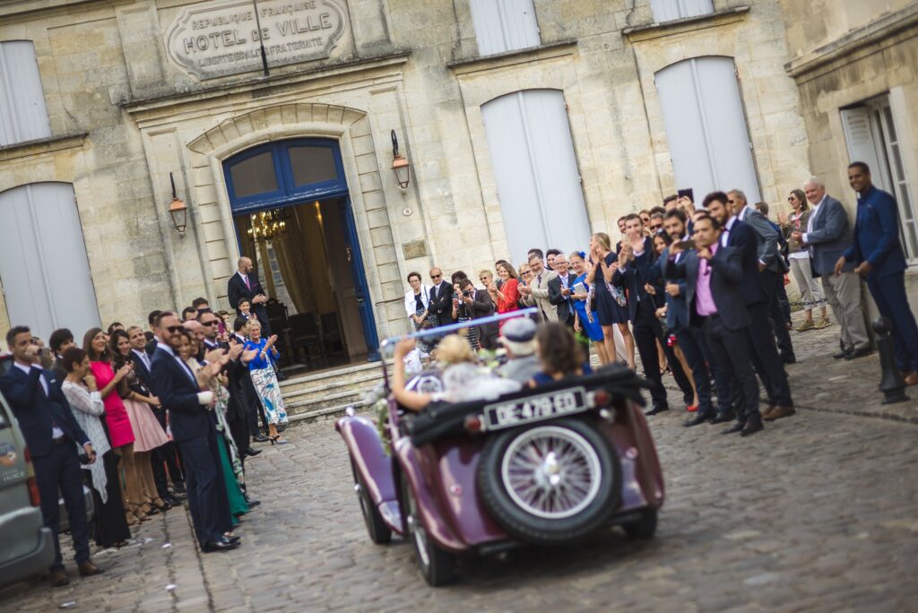 Une voiture ancienne ornée de fleurs arrive devant la mairie, sous les regards d'invités rassemblés.