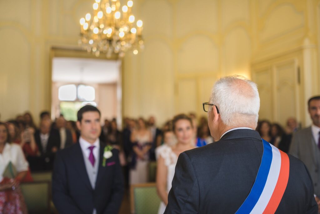 Dans la salle de mairie, un marié écoute un élu portant l'écharpe tricolore, devant les invités.