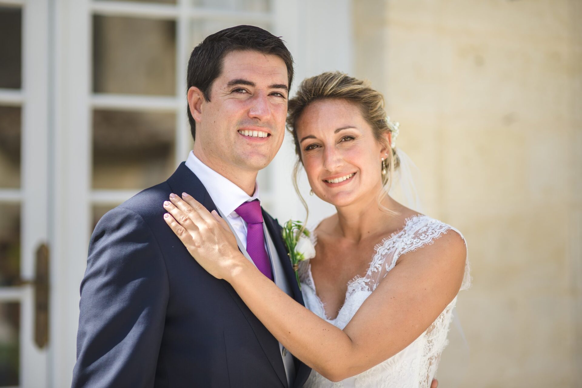 Un couple de mariés, joue contre joue, sourit devant une porte-fenêtre blanche.