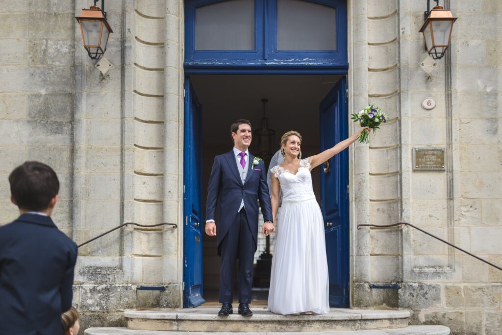 Les mariés sortent par une grande porte bleue, la mariée levant son bouquet.