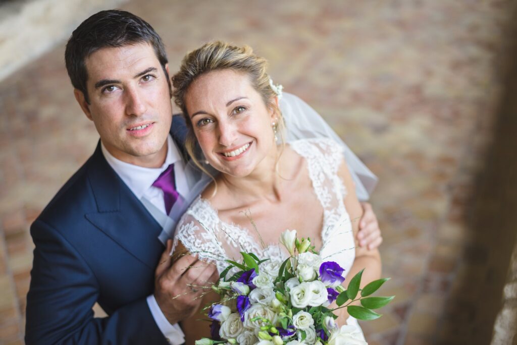 Portrait serré des mariés, enlacés et souriants, le bouquet de fleurs blanches et violettes au premier plan.