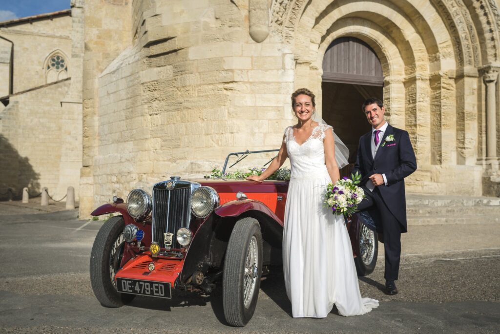 Les mariés posent près d'une voiture ancienne rouge, devant un portail de pierre.