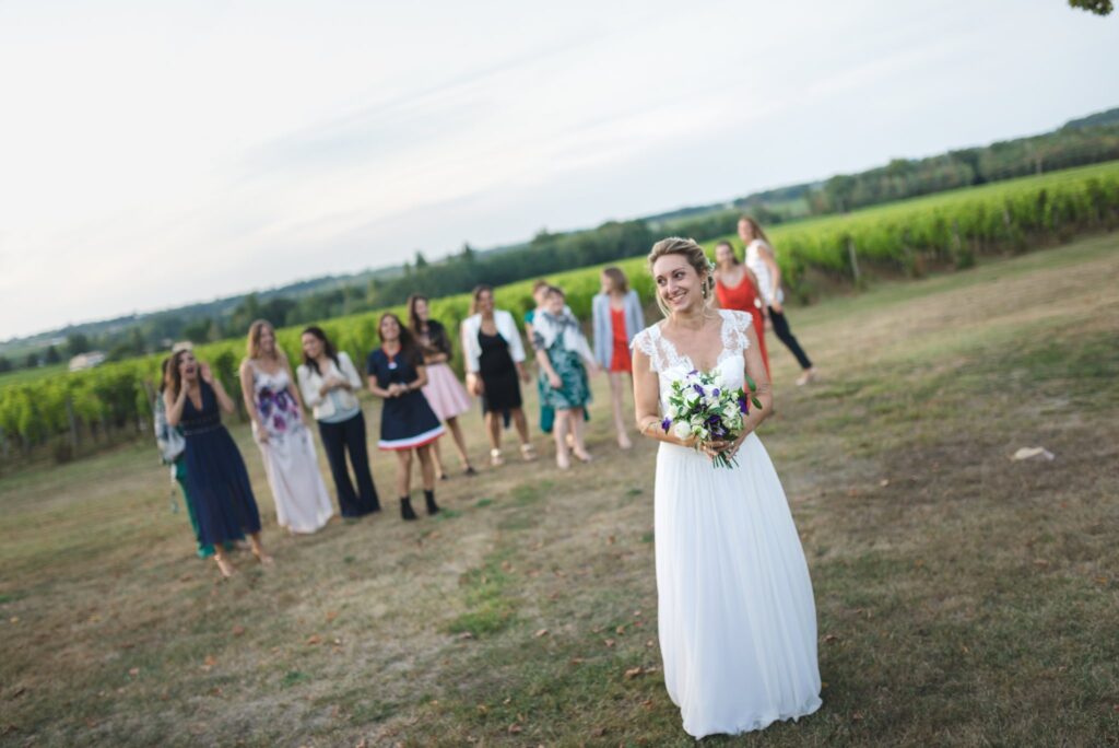Une mariée se tient bouquet en main, de dos à un groupe de femmes, sur une pelouse devant les vignes.