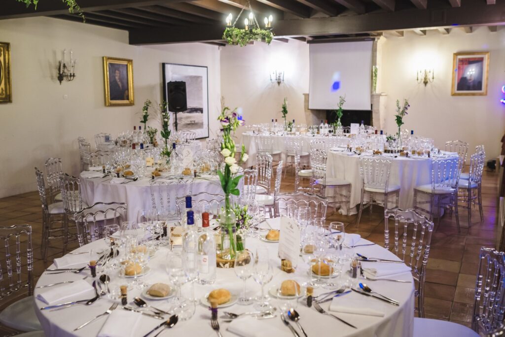 Une grande salle à poutres avec des tables rondes dressées pour le dîner.