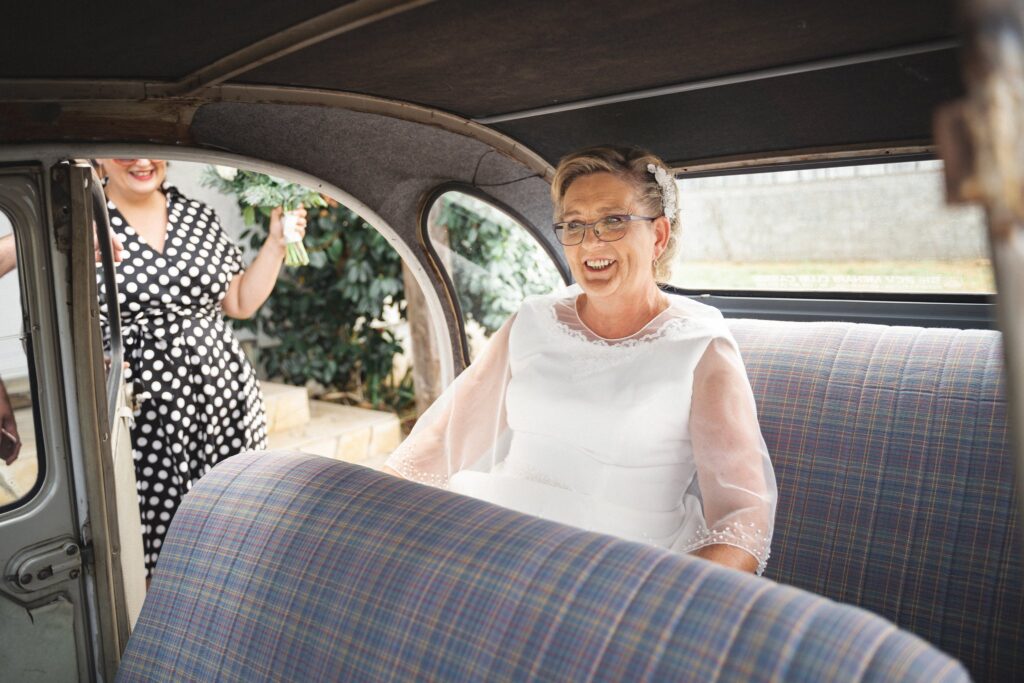 Une mariée souriante assise à l'arrière d'une voiture ancienne, une femme en robe à pois debout à la portière.