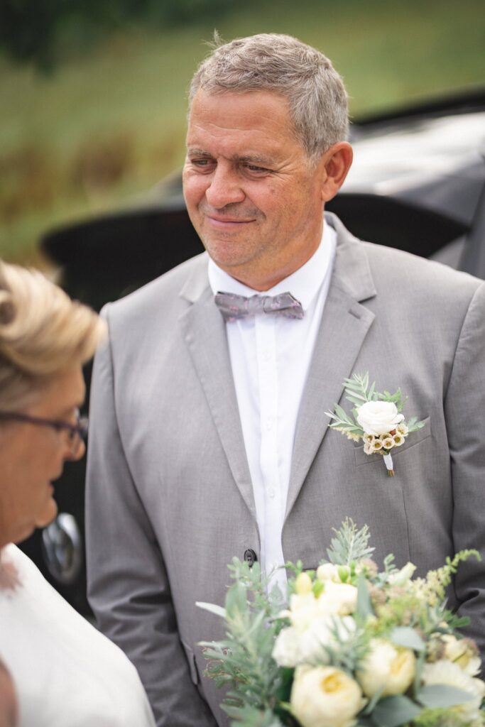 Un marié en costume gris et nœud papillon, souriant, un bouquet blanc au premier plan.