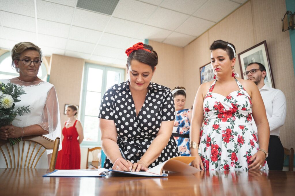 Une femme en robe à pois signe le registre dans une salle de mairie, entourée de plusieurs personnes.