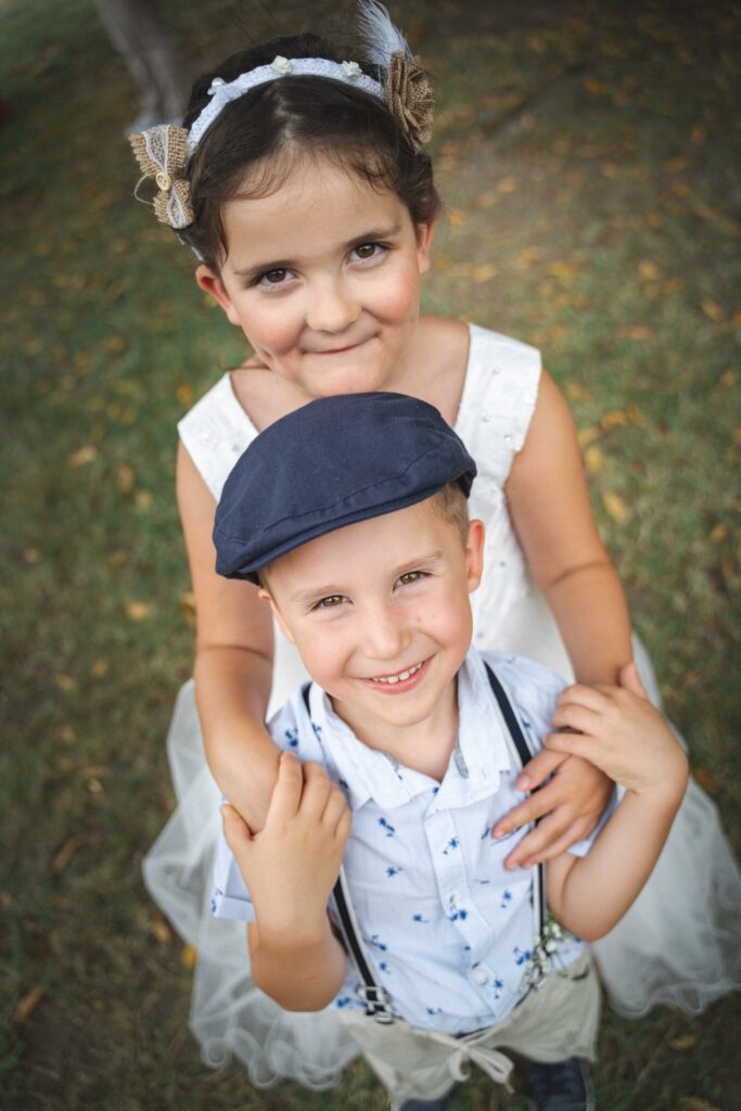 Une fillette en robe blanche et un garçon en casquette sourient à l'objectif, vus d'en haut.