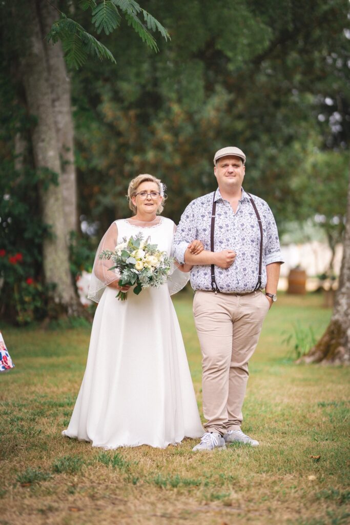 Une mariée avec son bouquet marche bras dessus bras dessous avec un homme en casquette et bretelles sur une pelouse.