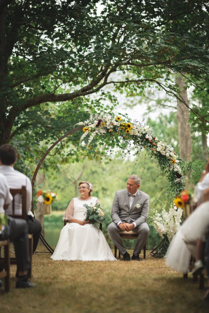 Les mariés assis sous une arche de fleurs, face à des invités, entre de grands arbres.