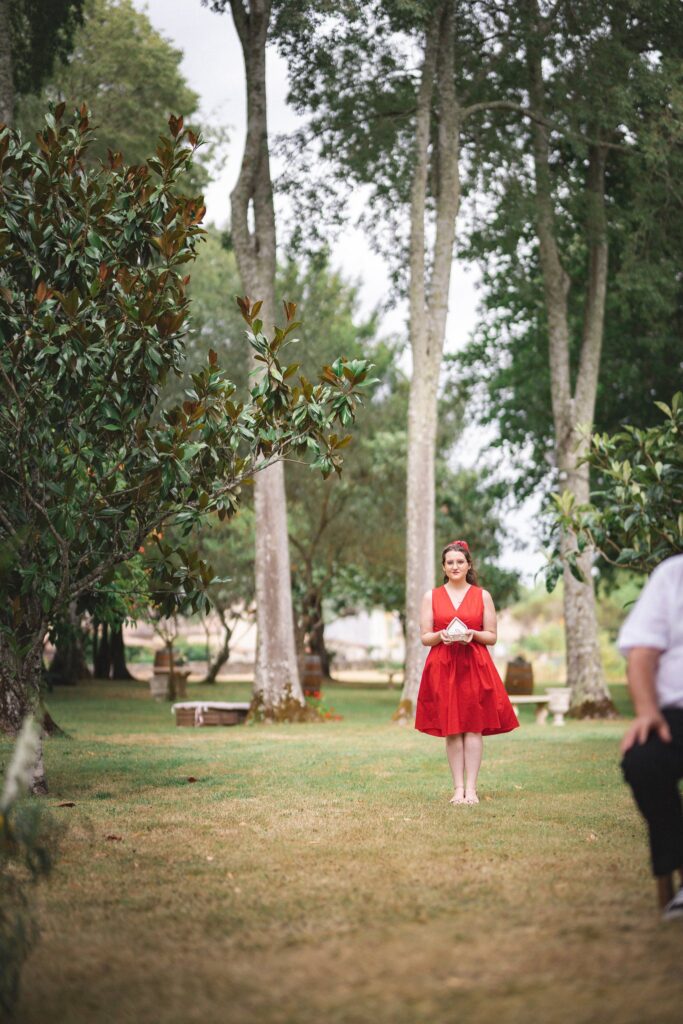 Une femme en robe rouge debout sur l'herbe entre de hauts troncs d'arbres.